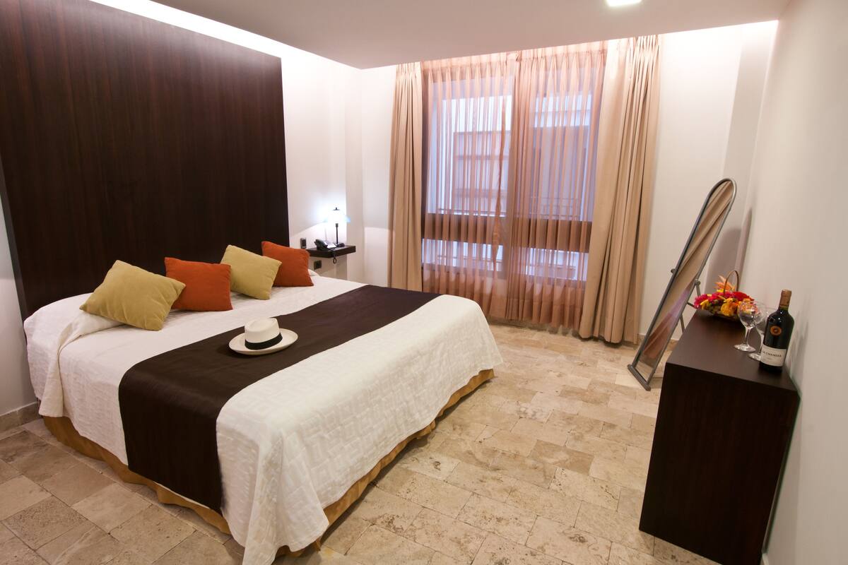 Habitación de hotel con una cama grande, una mesita de noche con lámpara, un espejo y un aparador con una botella y flores.