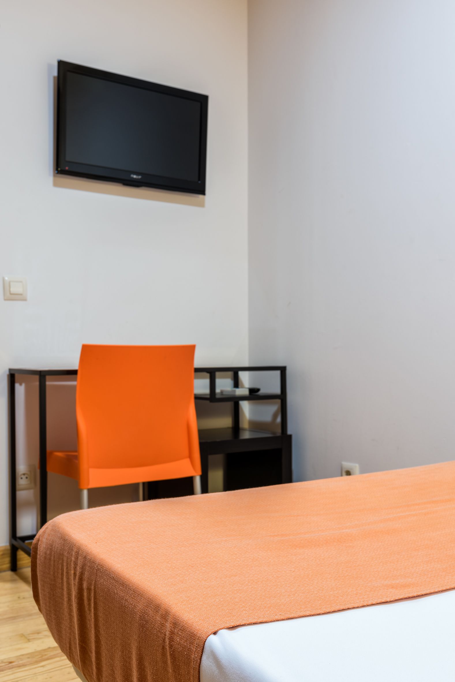 Een kamer met een bed, een oranje stoel, een bureau en een wandtelevisie.