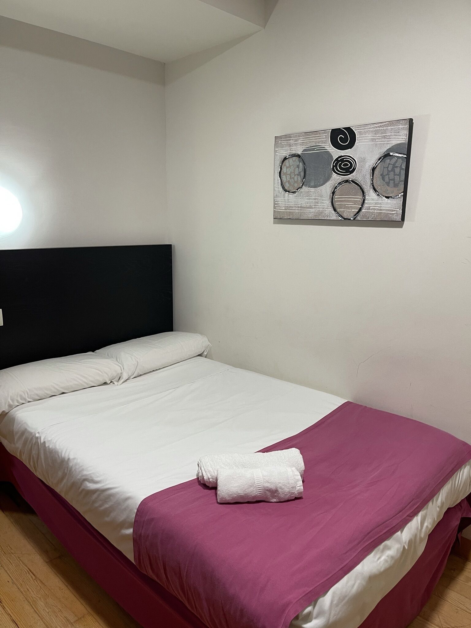 A single bed with a white and pink bedspread, a black headboard, and a wall-mounted abstract art piece.