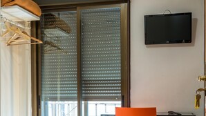 Quarto de hotel com uma cama, uma poltrona laranja, uma televisão na parede e uma porta de correr para a varanda.