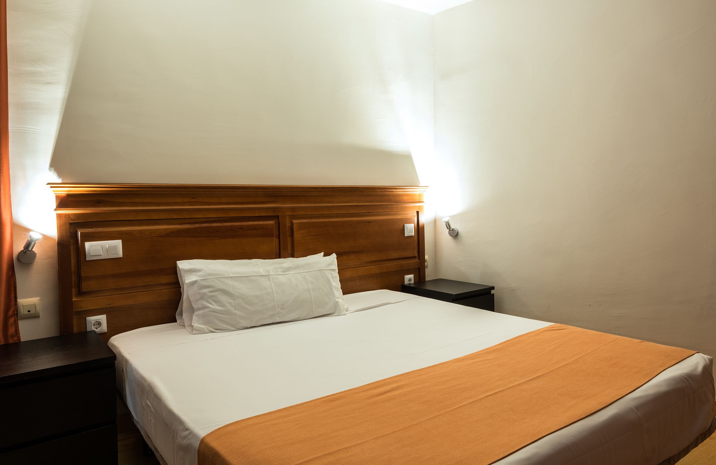 Habitación de hotel con cabecera de madera, cubrecama blanco y manta color naranja.