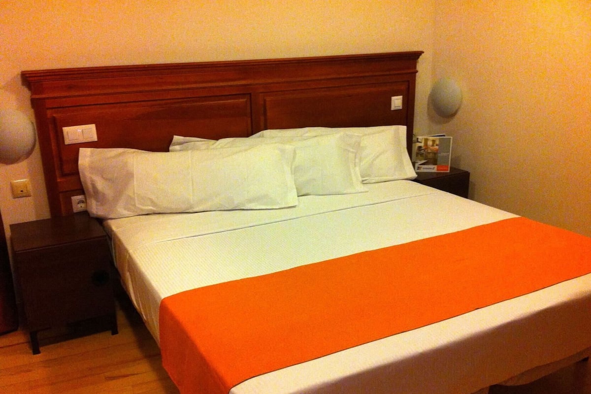 Una habitación de hotel estándar con cabecera de madera, una cama individual con ropa blanca, una manta naranja, una mesita de noche con lámpara y una pequeña mesa con un libro.