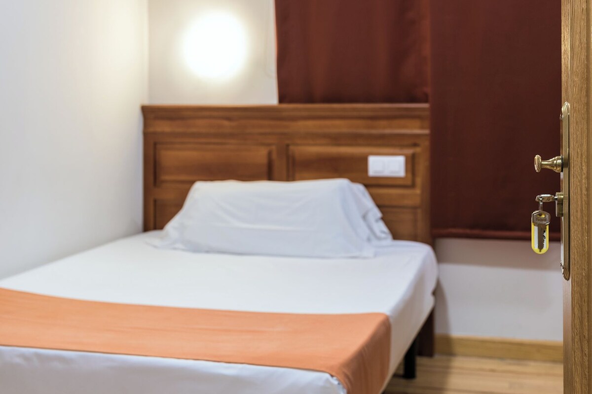 A hotel room with a wooden bed, white bedding, a wooden headboard, a brown curtain, and a wooden floor.