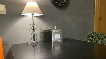 Una mesa con una lámpara, un cubo de basura, un código QR para autoservicio y un reloj en la pared.