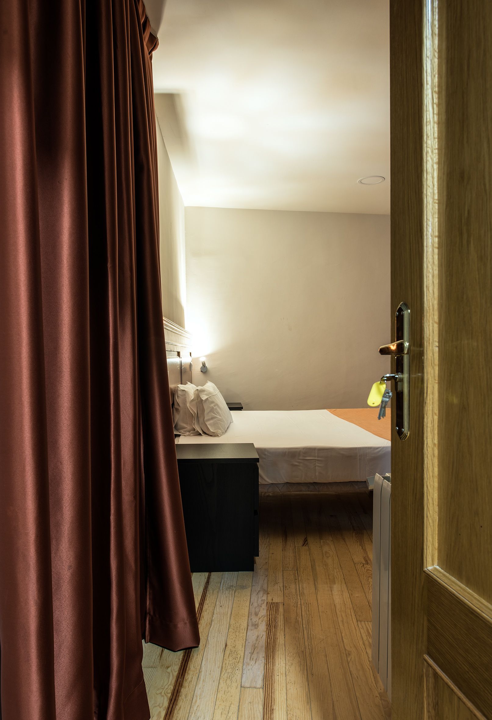 Ein kleines Zimmer mit einem Einzelbett, Holzboden, Bettwäsche in Weiß, einem Nachttisch und einer Tür mit Griff.