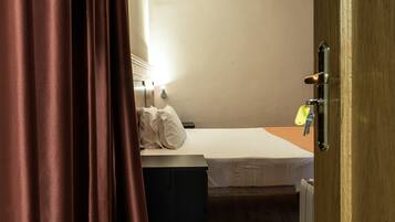 Een kleine kamer met een enkel bed, houten vloer, nachtkastje en een deur met een hendel. Beddengoed is wit.