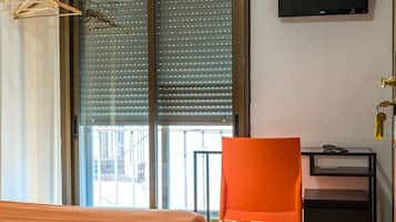 Habitación de hotel con cama, una butaca naranja, escritorio, televisor y un ventanal con persianas.