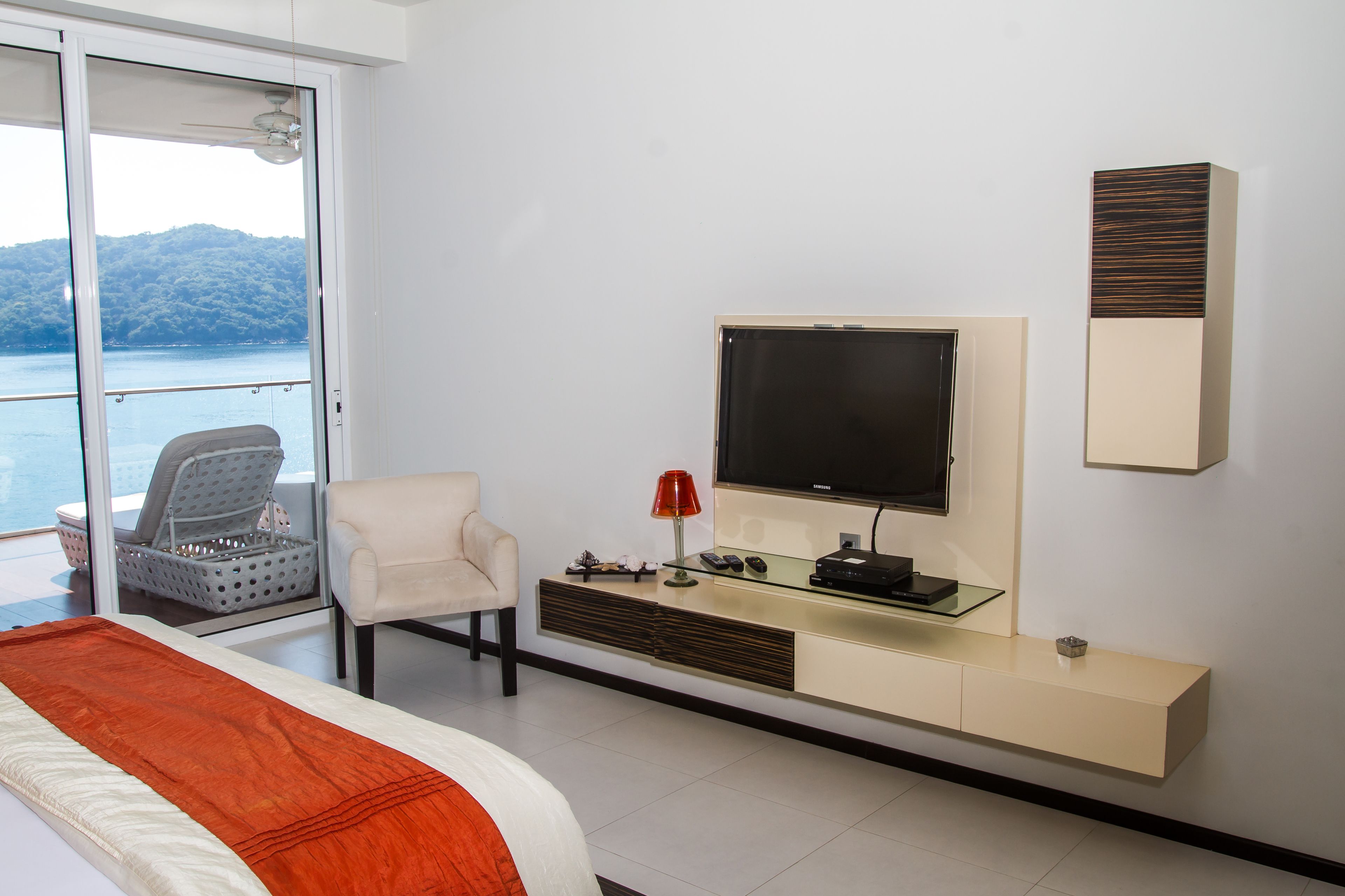 Una habitación de hotel moderna con una cama grande, televisión, zona de estar con una silla y vista al mar a través de una puerta corrediza.