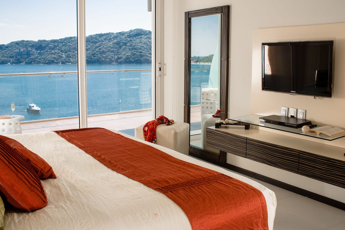 A hotel room with a large bed, a TV, a glass table, and a view of a body of water.
