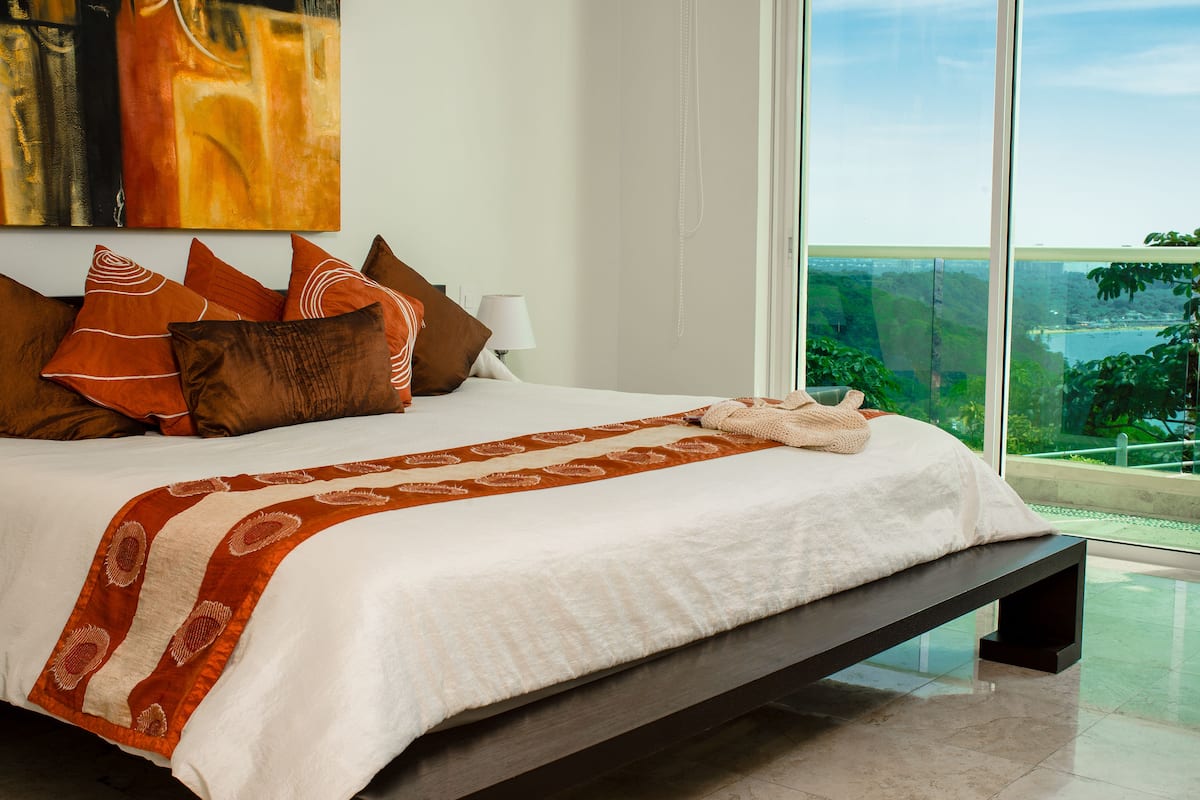 A modern bedroom with a large bed, a view of greenery, and a painting on the wall.
