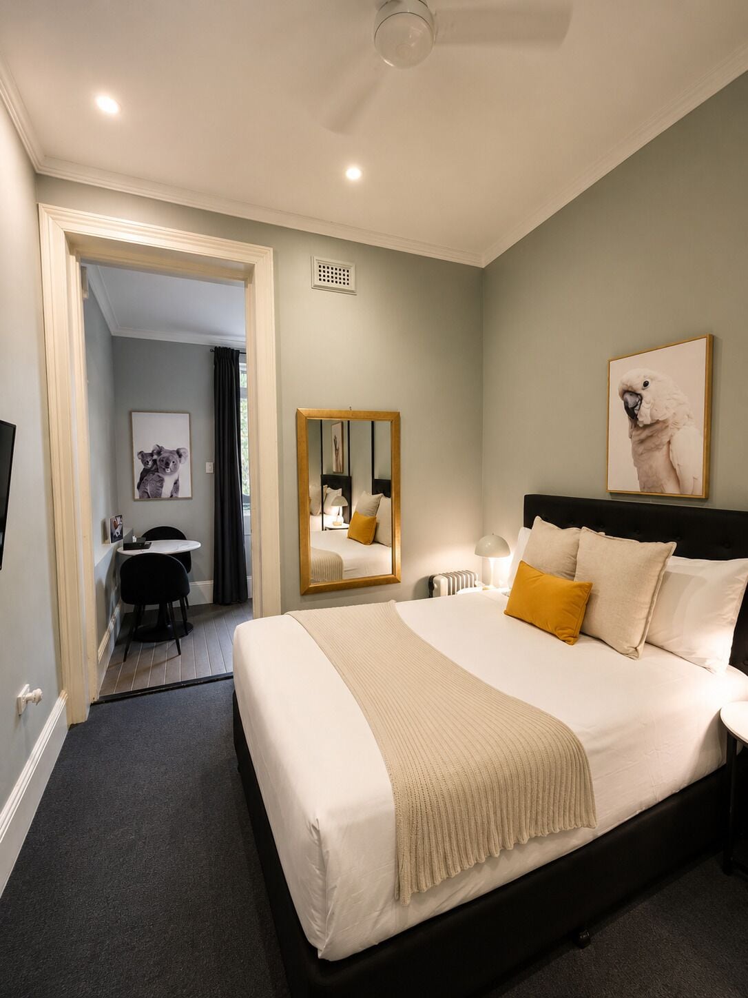 A hotel room with a bed, bedside table, mirror, artwork, and a desk with a chair.