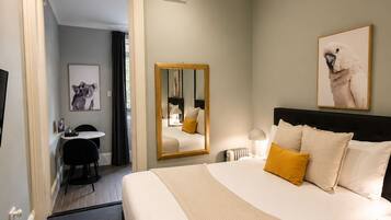 A hotel room with a bed, bedside table, mirror, artwork, and a desk with a chair.