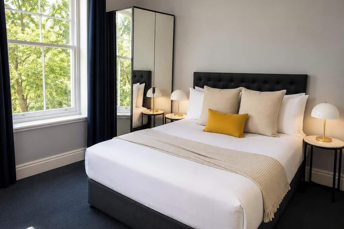 Une chambre d’hôtel avec un grand lit, un miroir, deux tables de chevet avec des lampes et une vue sur les arbres par la fenêtre.