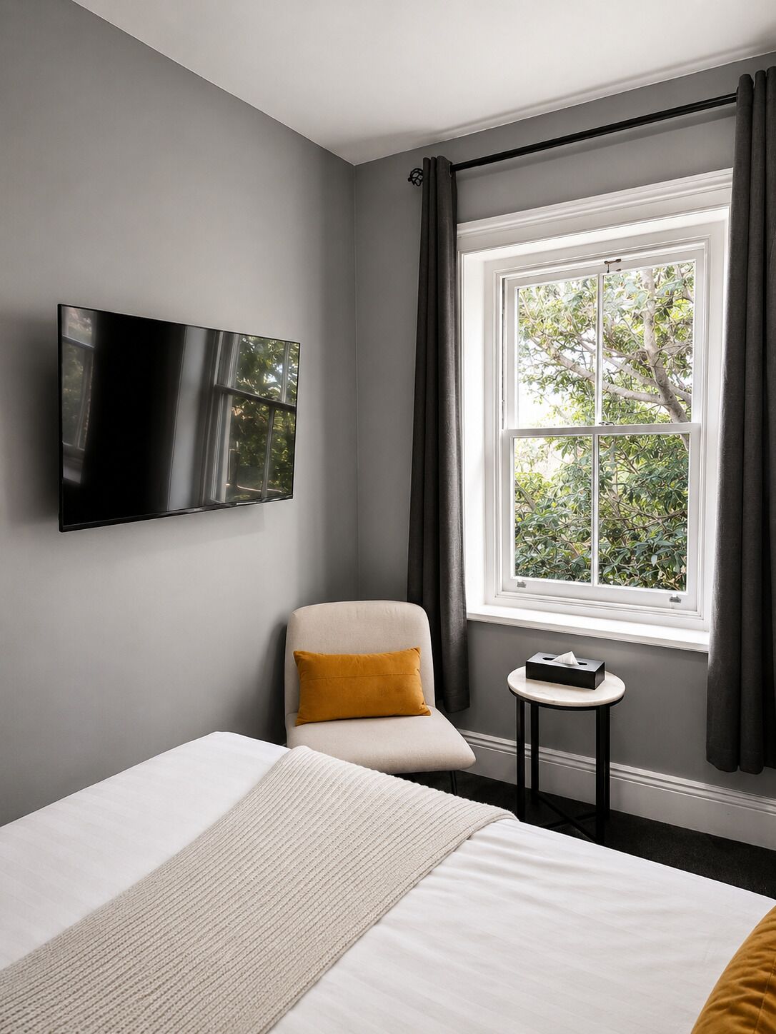 A modern hotel room with a bed, a television mounted on the wall, a small table with a tissue box, and a window with curtains.