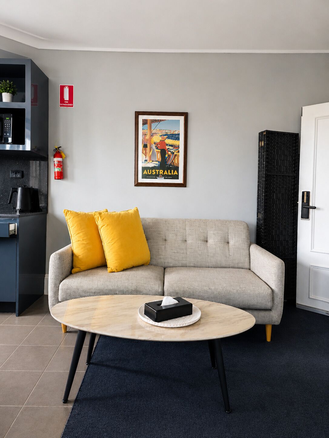 Ein Wohnzimmer mit einer Couch, einem Couchtisch, einem gerahmten Bild, einem Feuerlöscher und einer Küchenzeile.