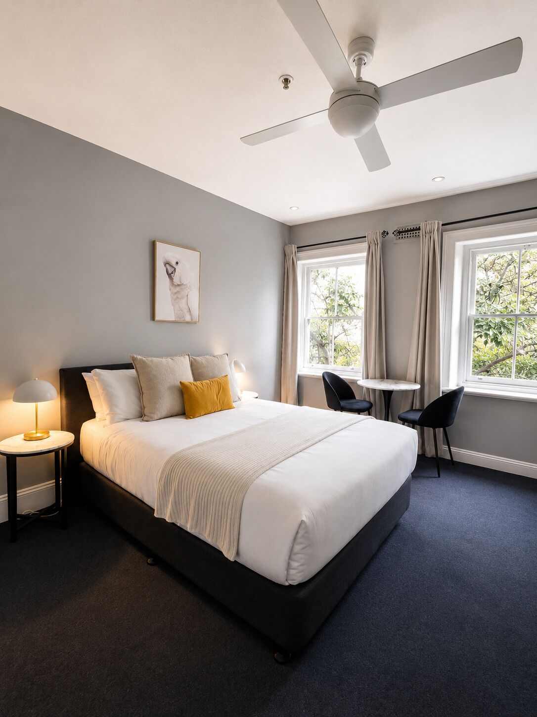 A modern hotel room with a large bed, a ceiling fan, a small table with chairs, and a window with curtains.