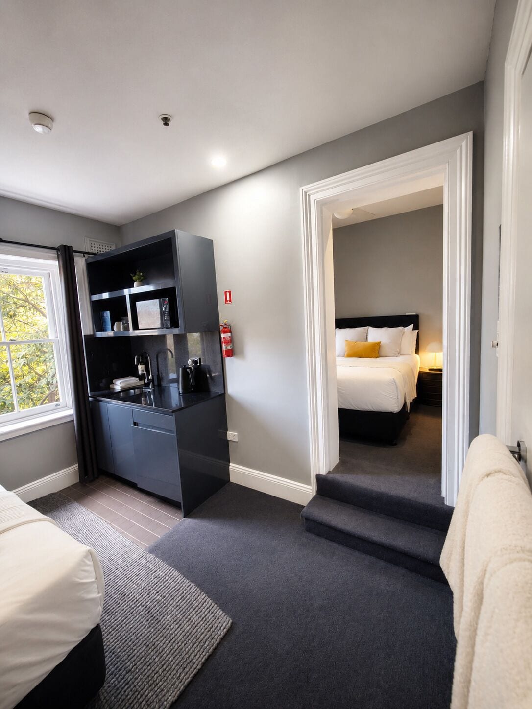 A modern hotel room with a kitchenette, a bed with white bedding, a grey carpet, and a view of greenery outside.