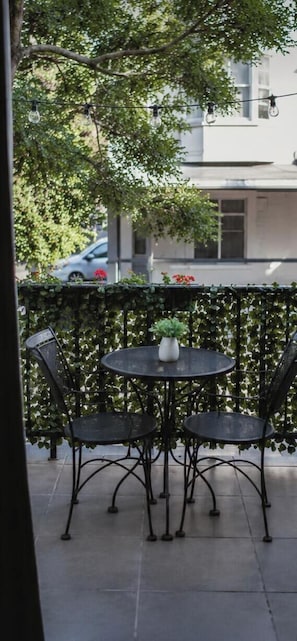 Un balcon avec une table ronde et quatre chaises, entouré d’une clôture en métal noir ornée de plantes grimpantes.