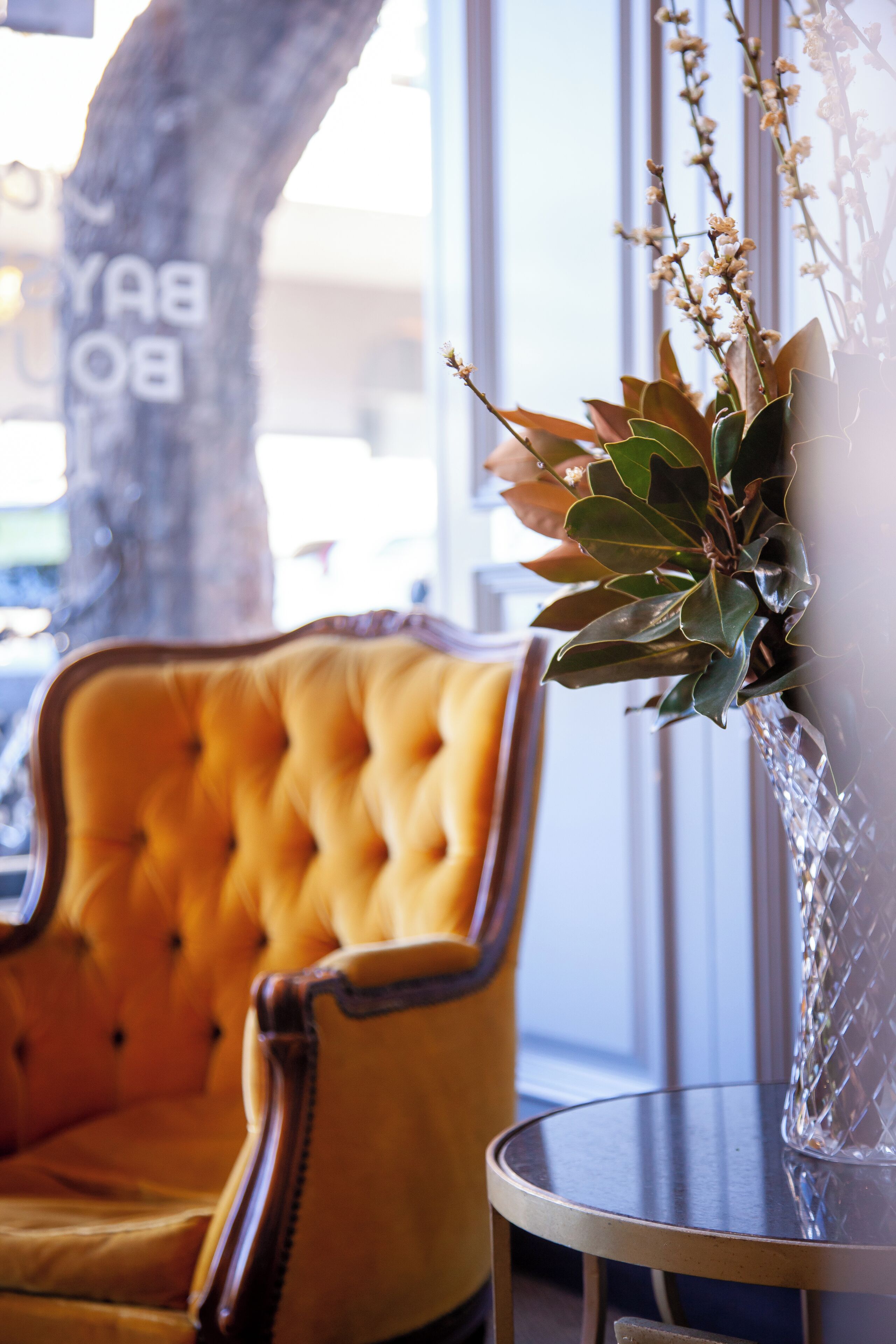 A yellow tufted chair, a glass vase with green foliage, and a tree with a sign reading "BAY BOY".
