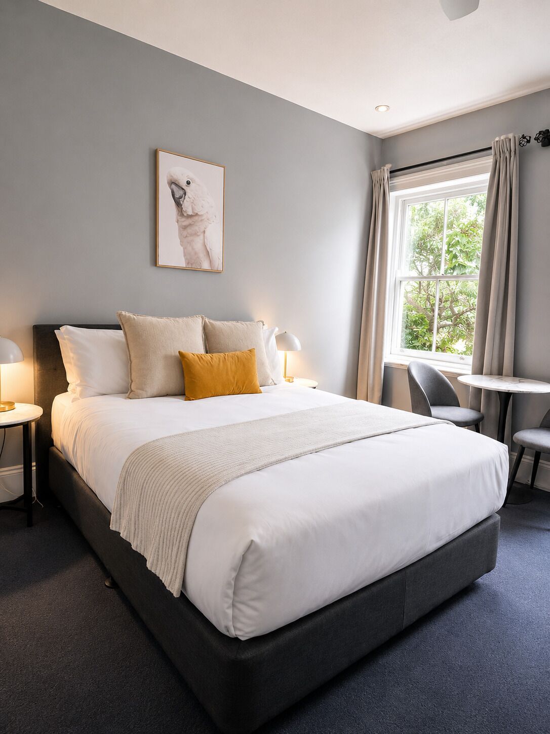 A modern hotel room with a large bed, bedside tables, a small round table, and a window with curtains.