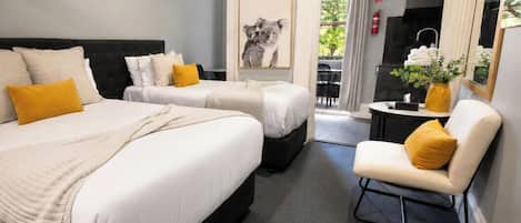 Hotellihuone, jossa on kaksi sänkyä, pieni oleskelualue, seinällä kehystetty taulu ja näkymä ulos.