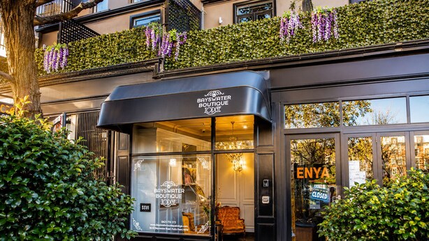 Entrada del hotel boutique Bayswater con fachada de vidrio, interior visible con un candelabro, y un letrero que indica que el hotel está cerrado.
