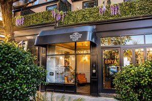 Entrada del hotel boutique Bayswater con fachada de vidrio, interior visible con un candelabro, y un letrero que indica que el hotel está cerrado.
