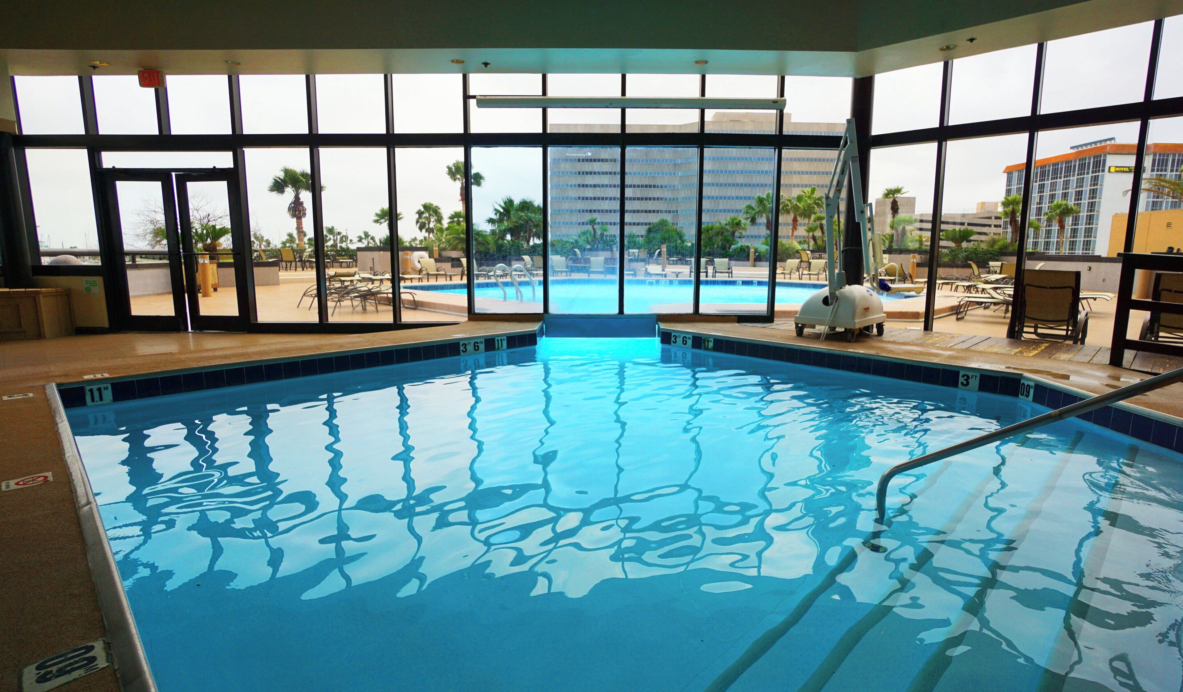 An indoor swimming pool with clear water, surrounded by lounge chairs and a view of palm trees and buildings.