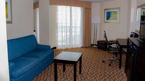Habitación de hotel con sofá azul, una mesita, un escritorio con computadora, una silla y un ventanal con cortinas.