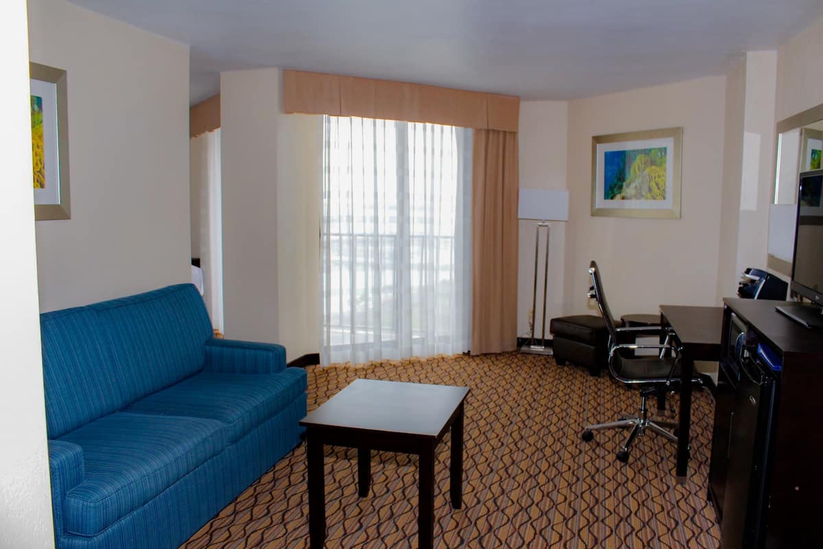 Ein Hotelzimmer mit blauem Sofa, einem kleinen Tisch, einem Schreibtisch mit Computer, einem Stuhl und einem großen Fenster mit Vorhängen.