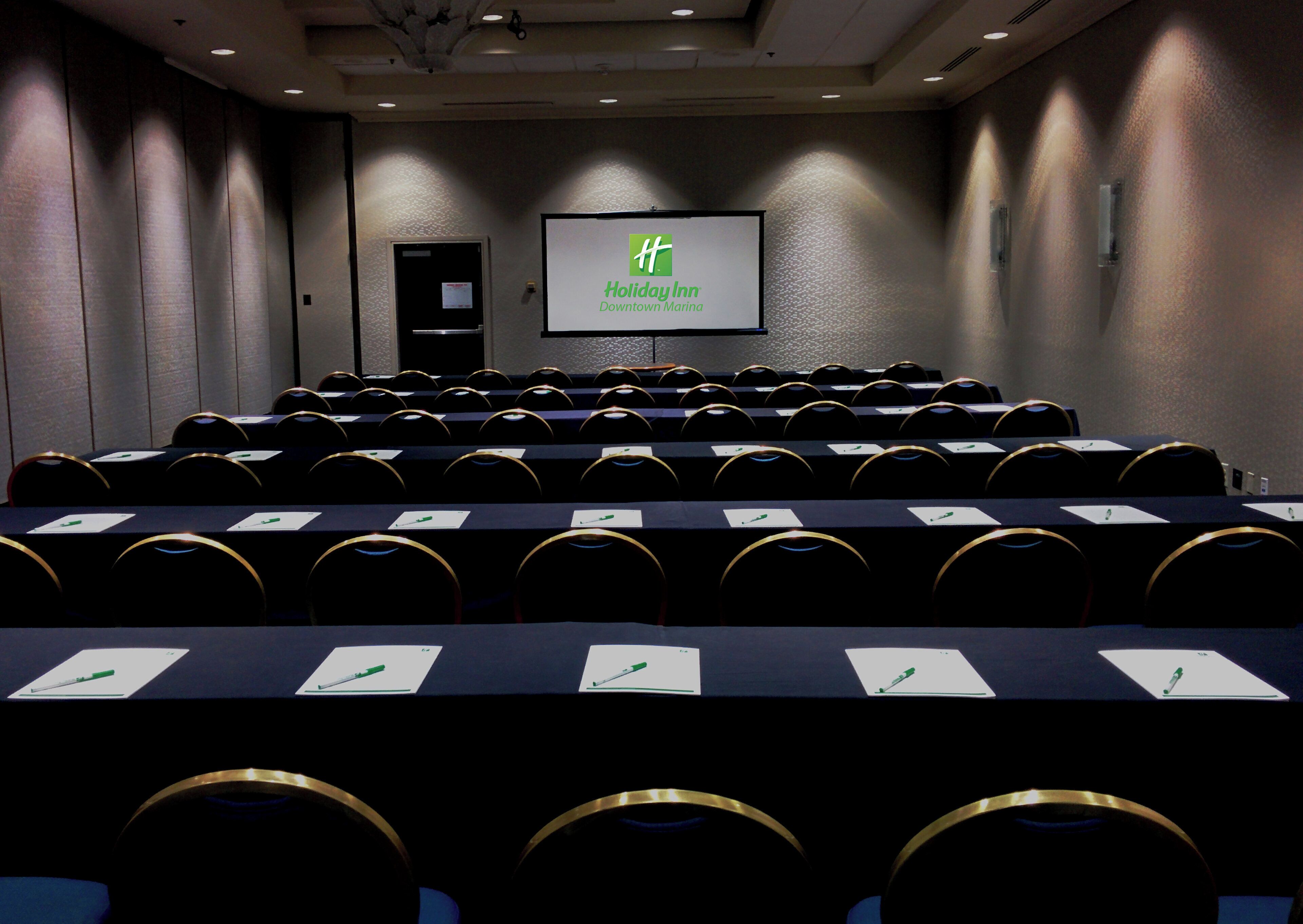 Un salón de conferencias con filas de mesas y sillas, una pantalla que muestra el logotipo de Holiday Inn y una puerta al fondo.