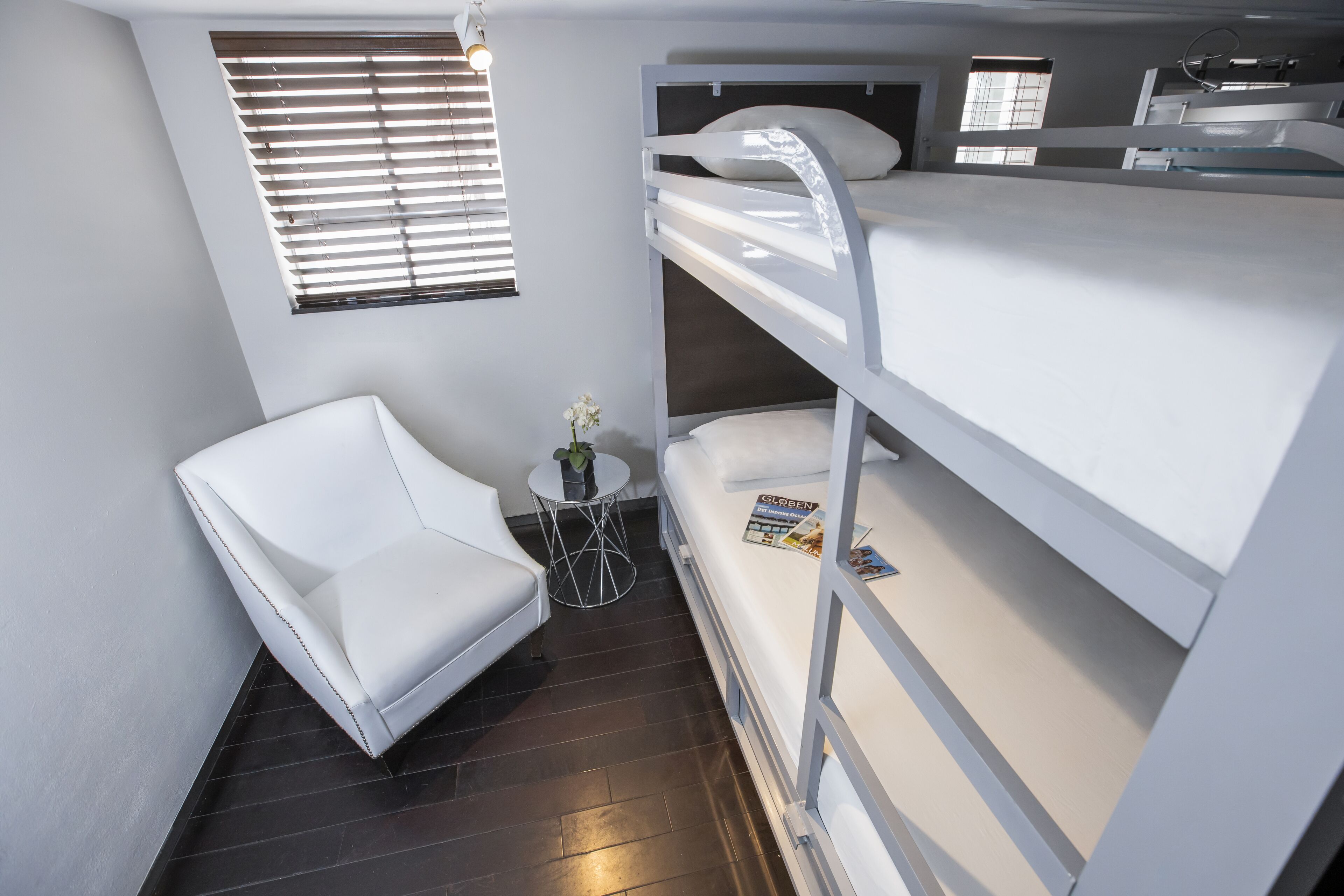A bunk bed room with a white chair, a small table with a plant, and a window with blinds.