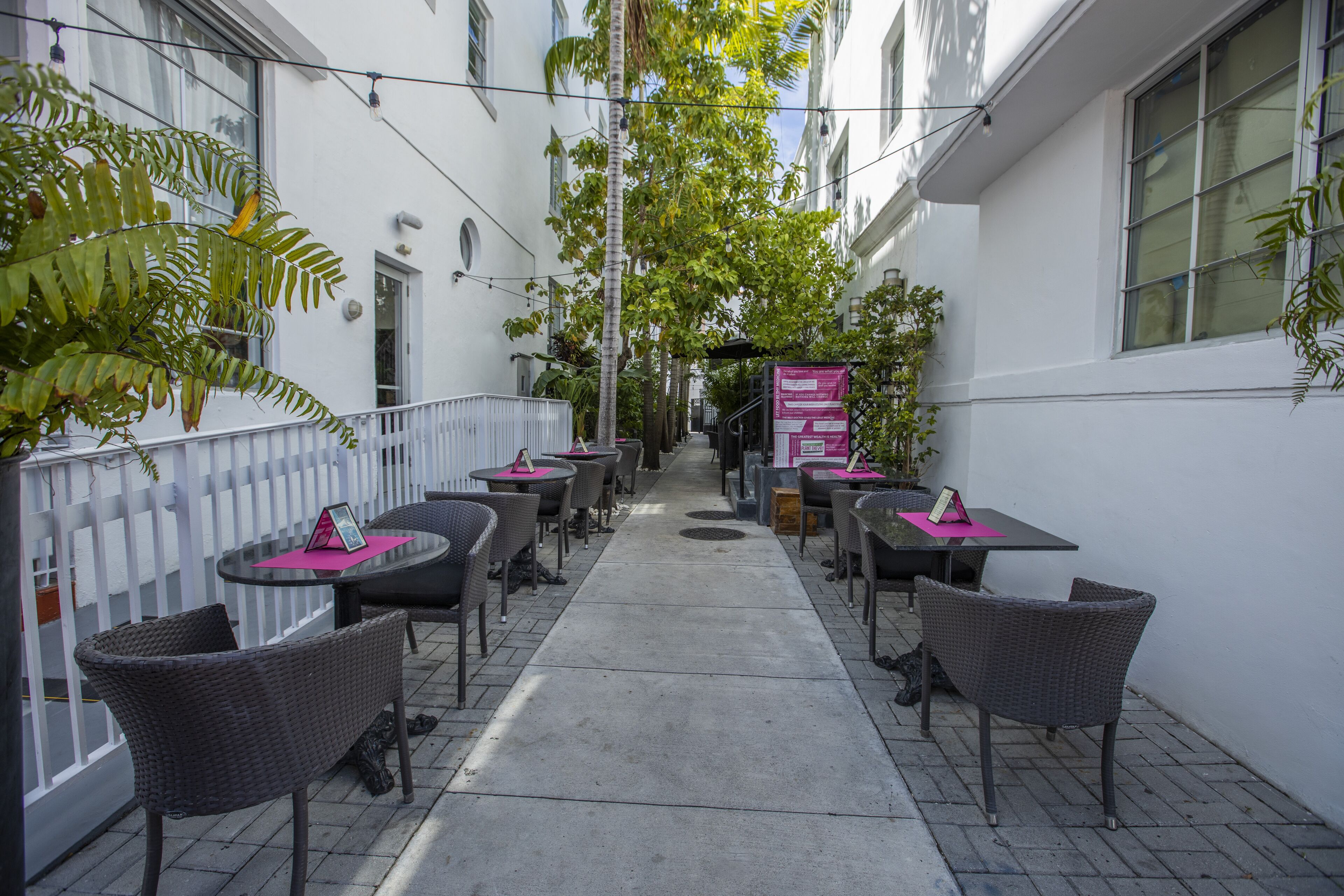 An outdoor cafe with wicker chairs and tables, a white fence, and a narrow pathway.