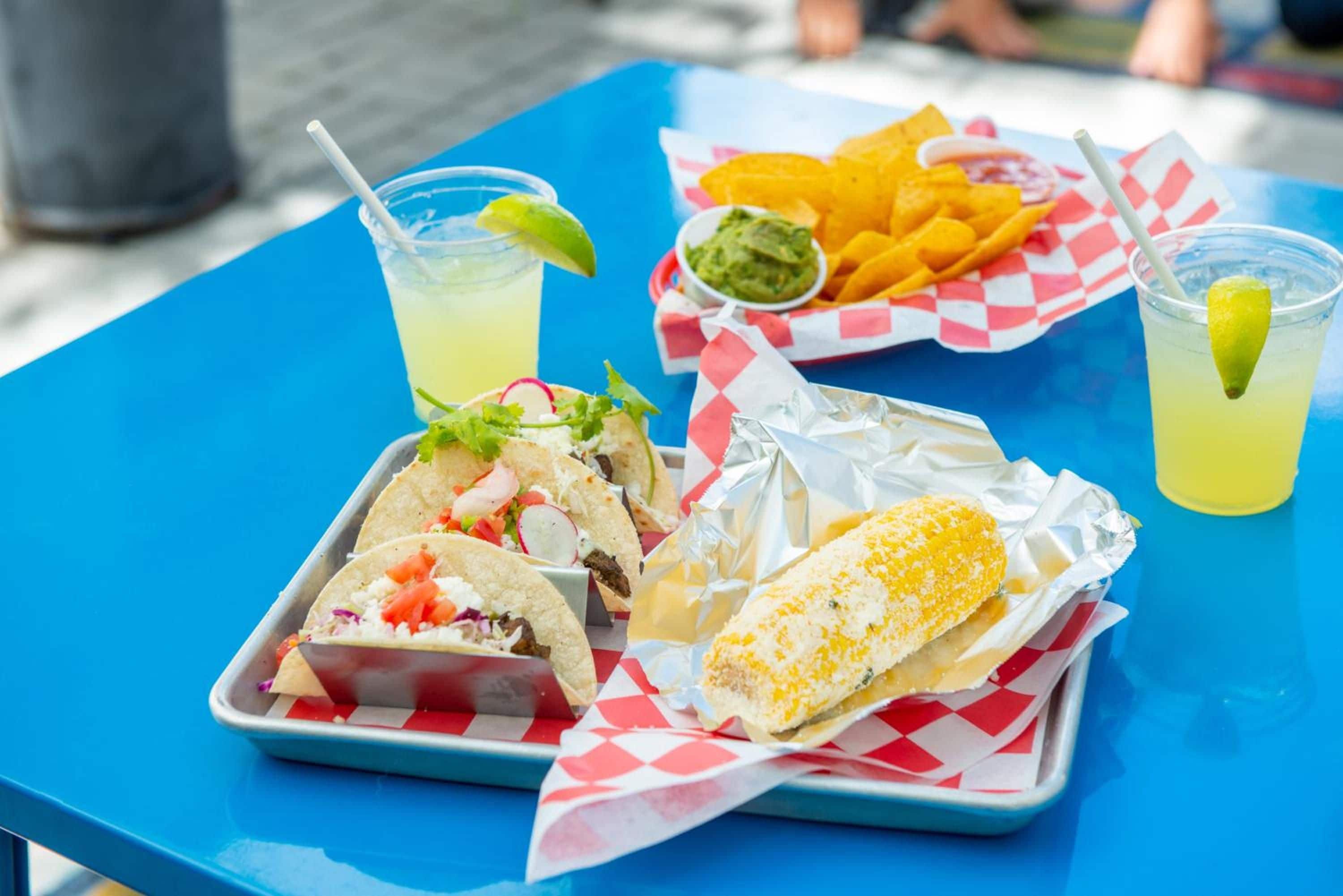 Une table bleue sur laquelle se trouve un plateau contenant des tacos, un épi de maïs et un bol de guacamole.