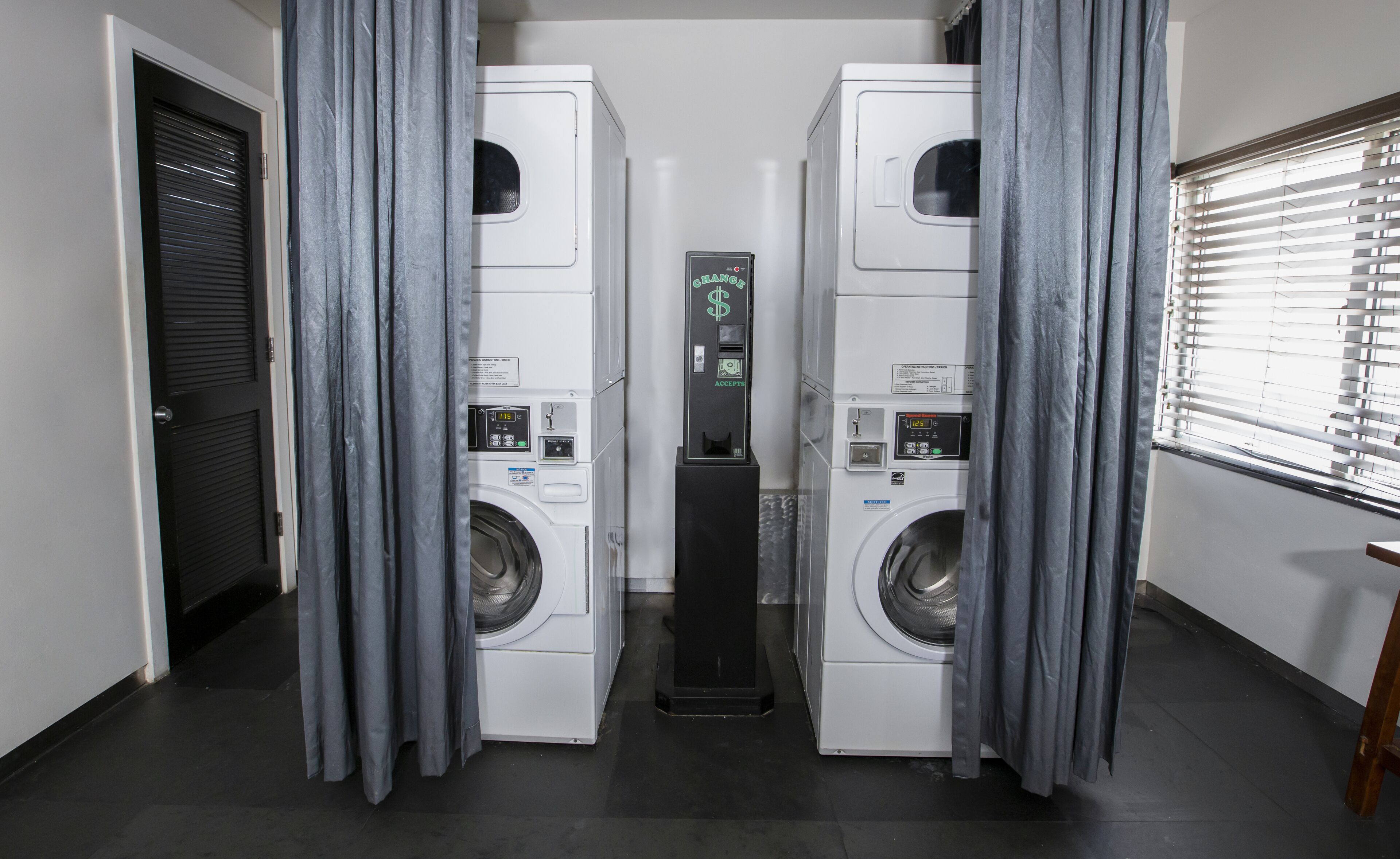 A laundry facility with two front-loading washing machines, a control panel, and grey curtains.