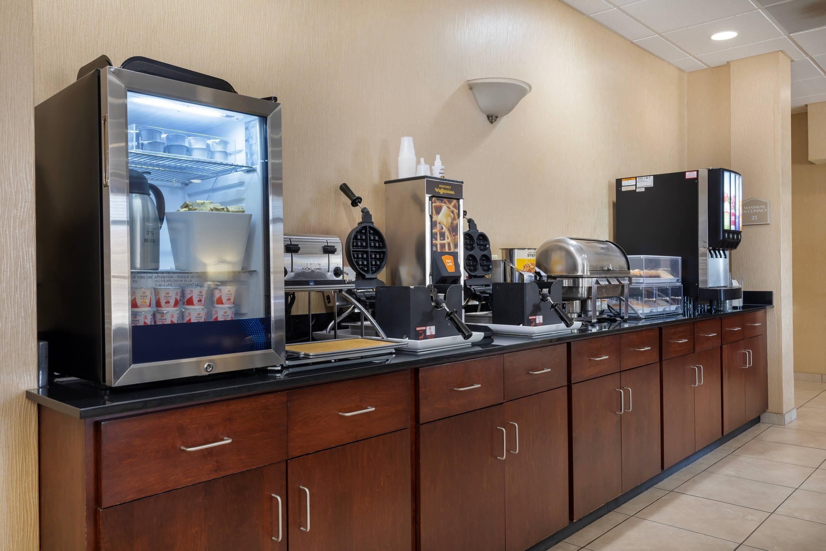 Un bar de desayuno autoservicio con refrigerador, máquina para gofres, cafetera y dispensador de bebidas.