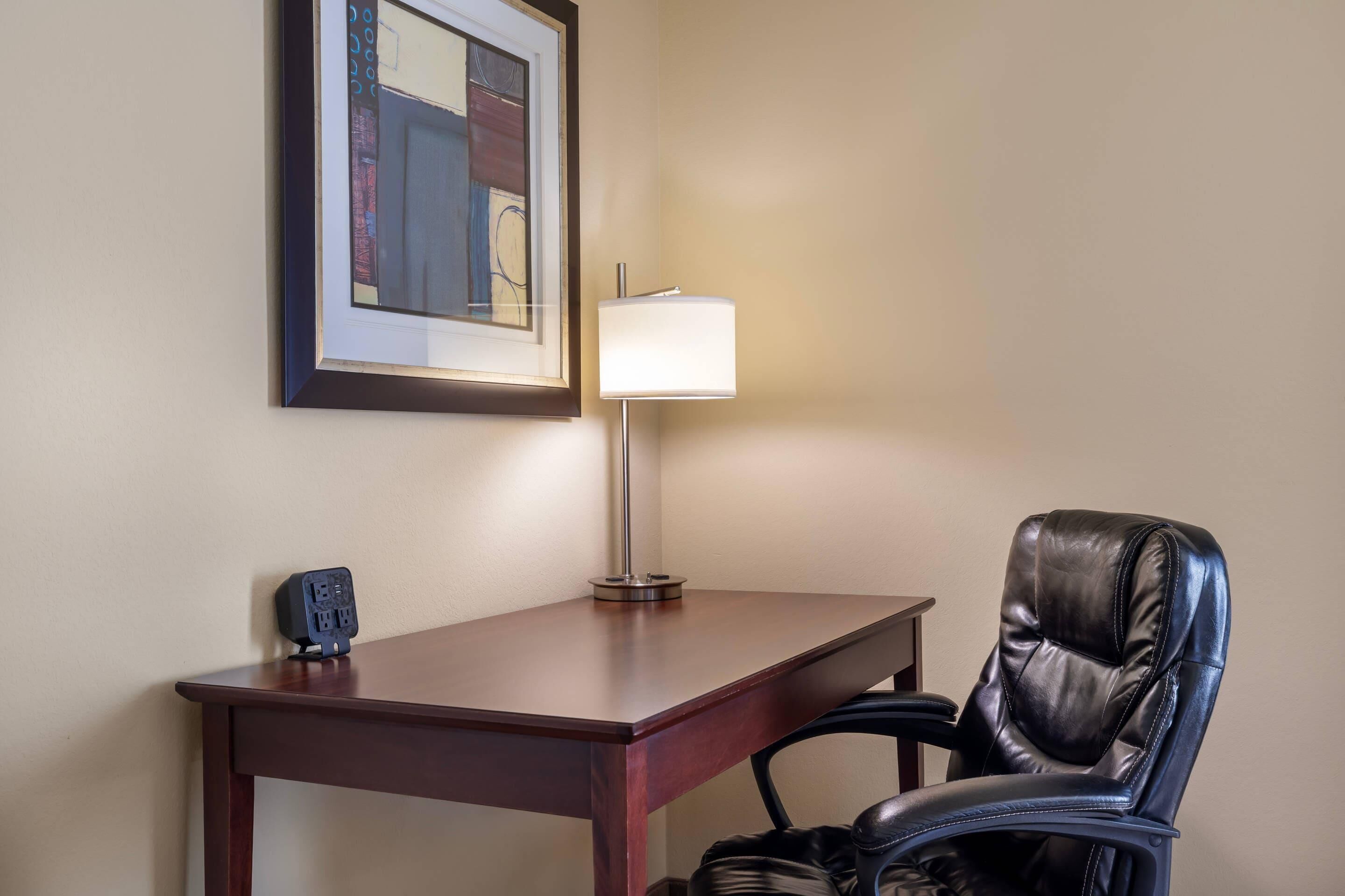 A hotel room with a desk, a chair, a lamp, and a framed picture on the wall.
