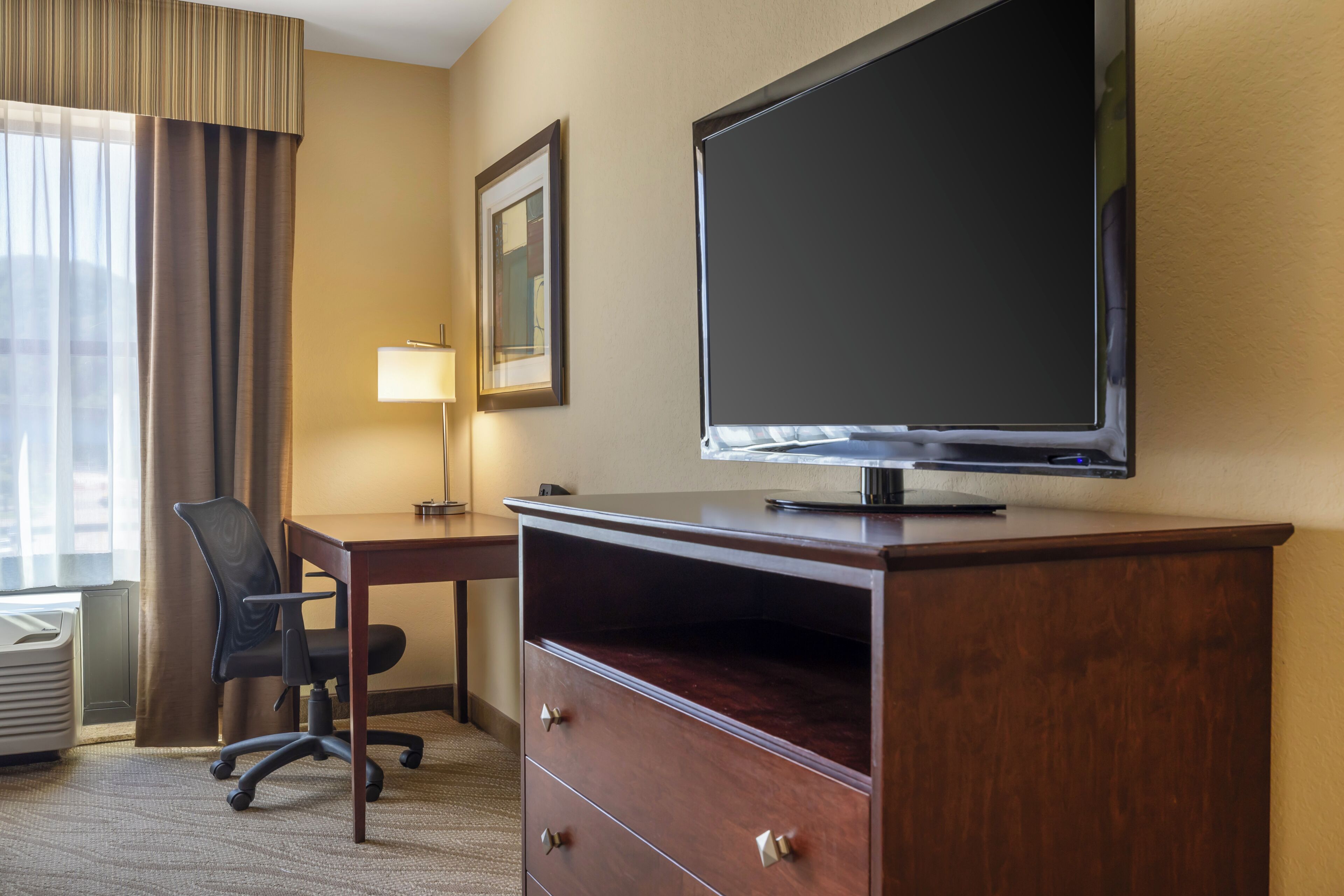 A hotel room with a flat-screen TV mounted on a wooden cabinet, a desk with a chair, a lamp, and a framed picture on the wall.