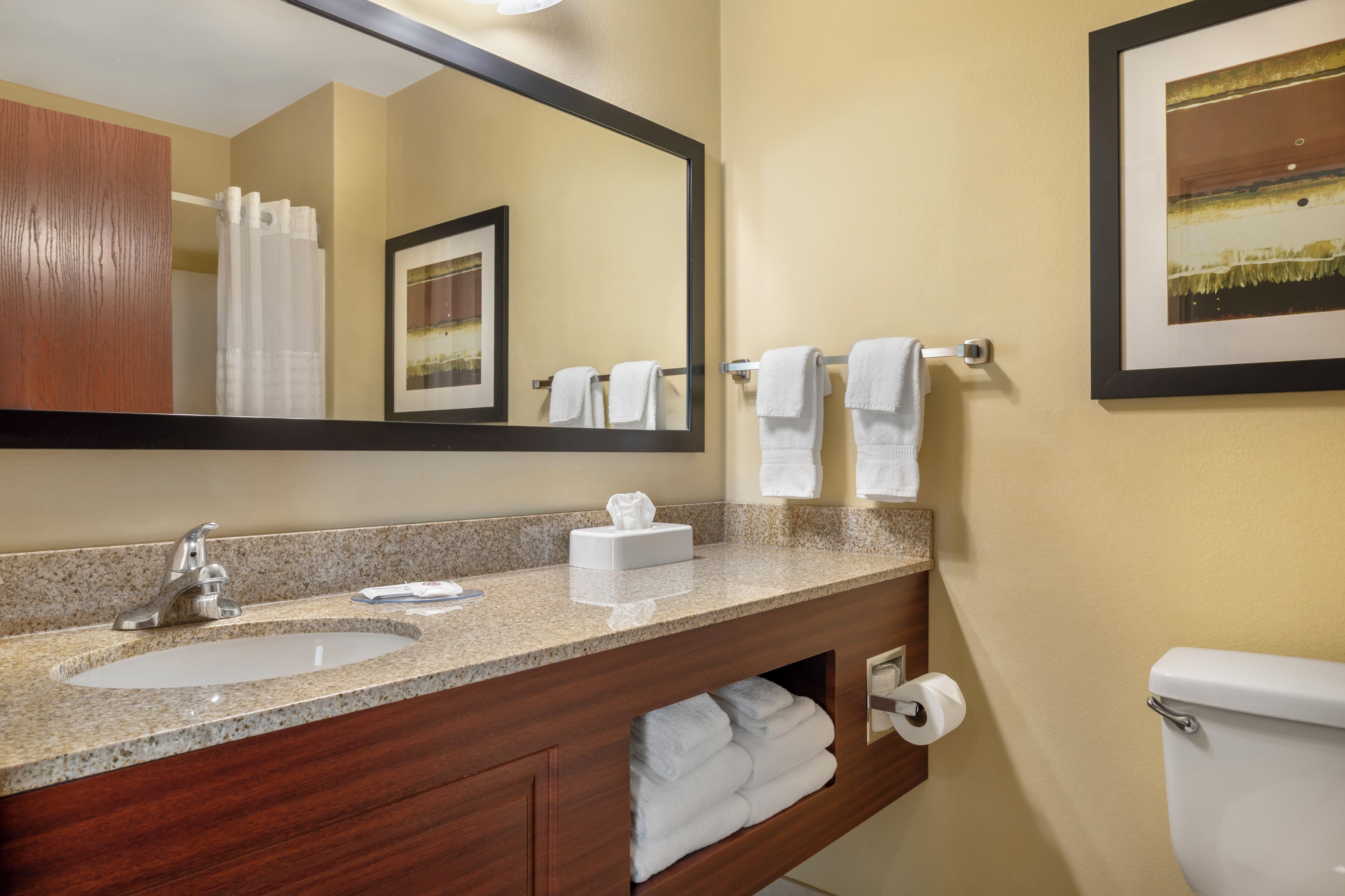 A bathroom with a granite countertop sink, a wooden vanity, a toilet, and a mirror with a framed picture.
