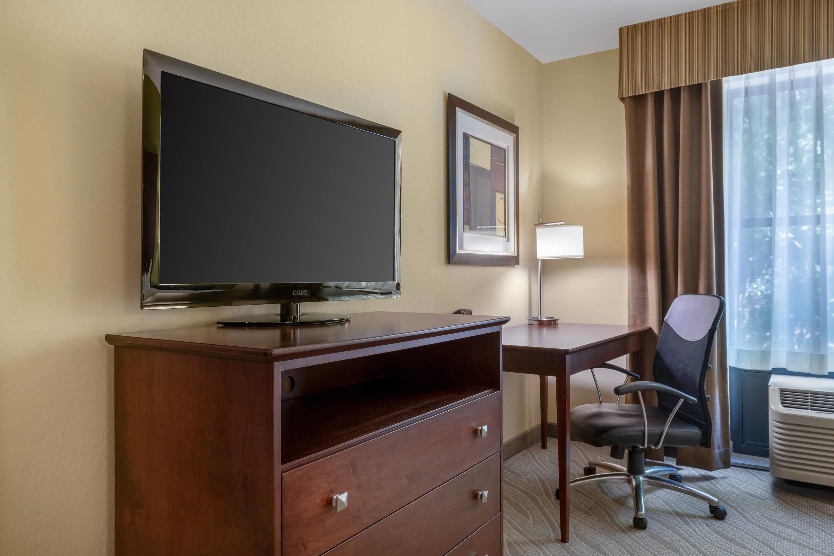 Habitación de hotel con televisión de pantalla plana, escritorio, silla, lámpara y ventana con cortinas.