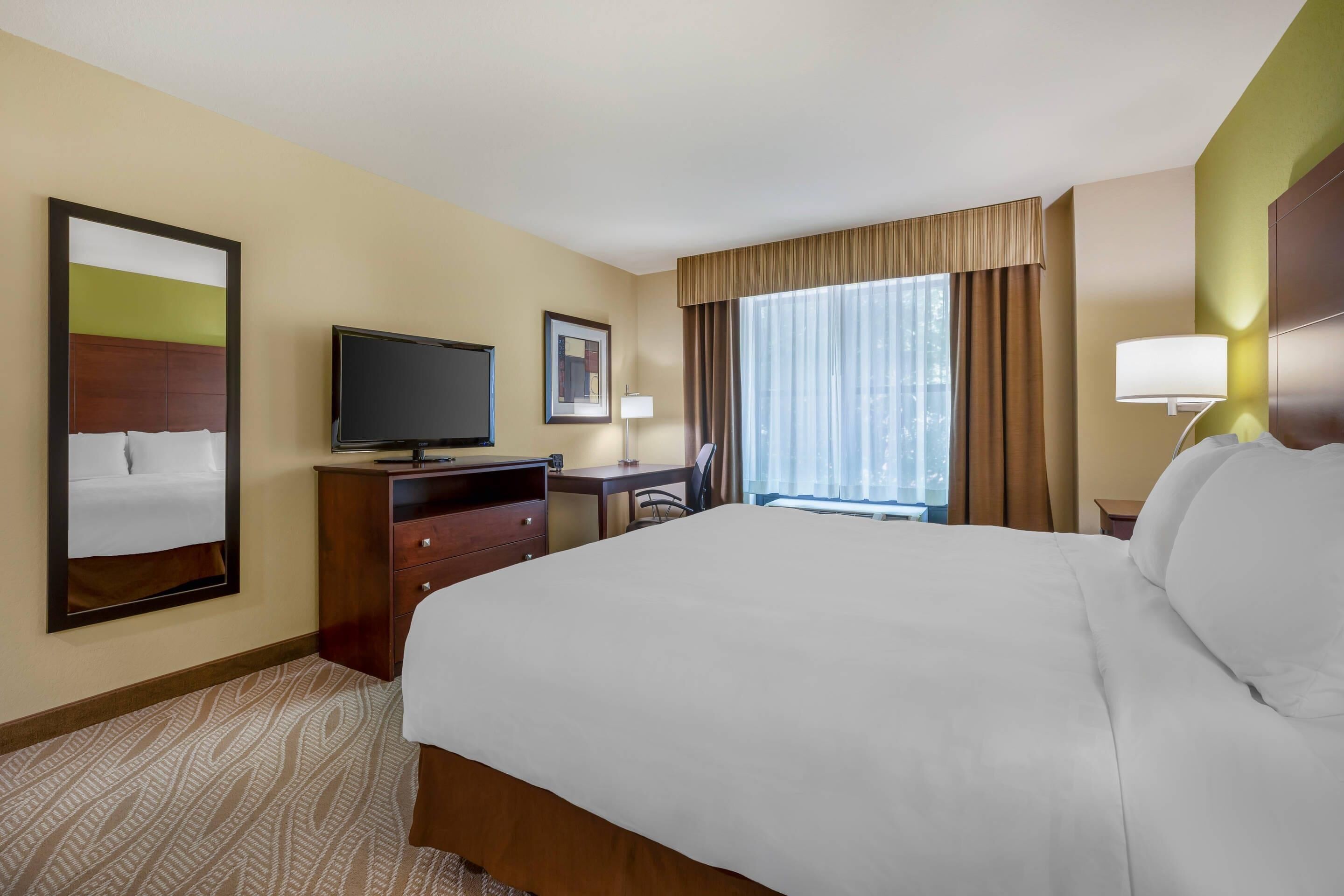 A hotel room with a large bed, a desk with a flat-screen TV, a chair, a lamp, and a window with curtains.