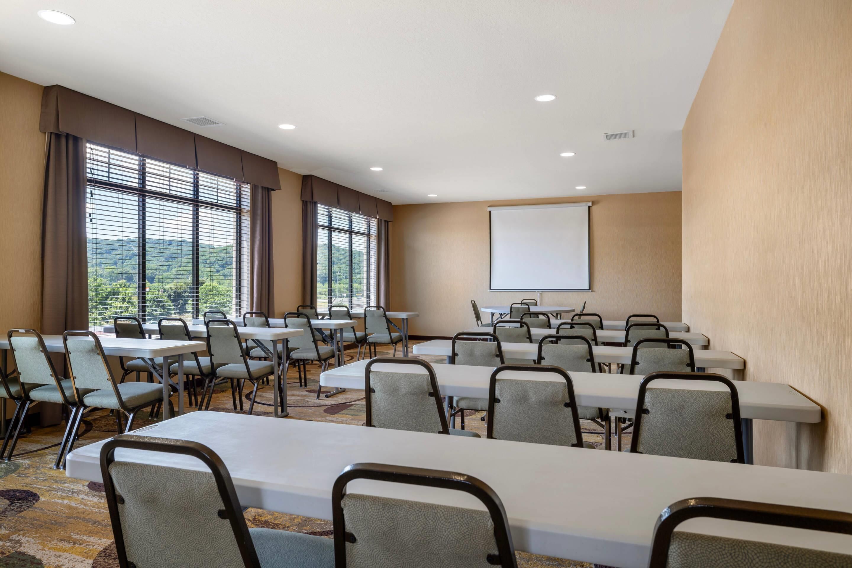 A conference room with multiple tables and chairs, large windows with blinds, and a projection screen on the wall.