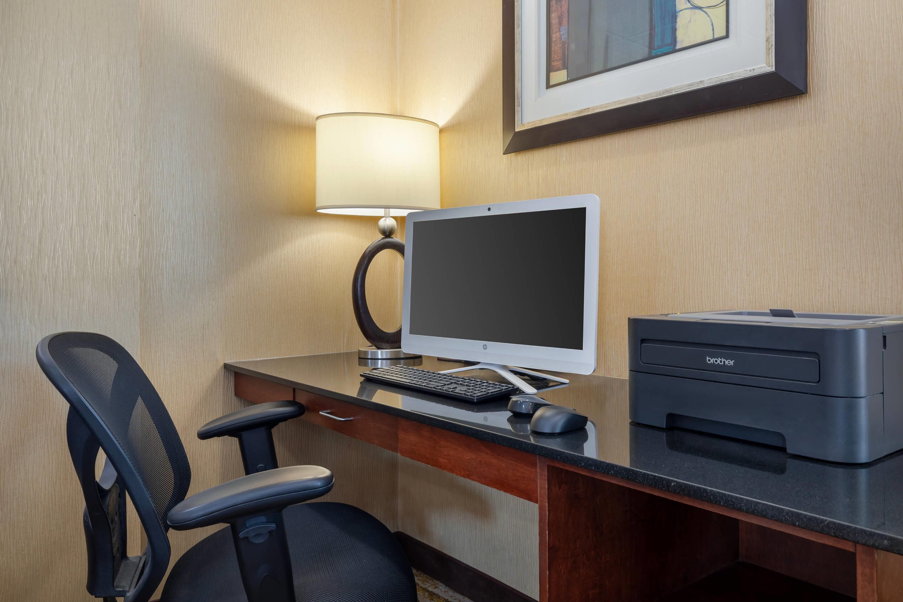 A hotel room desk with a desktop computer, a printer, a chair, and a lamp.