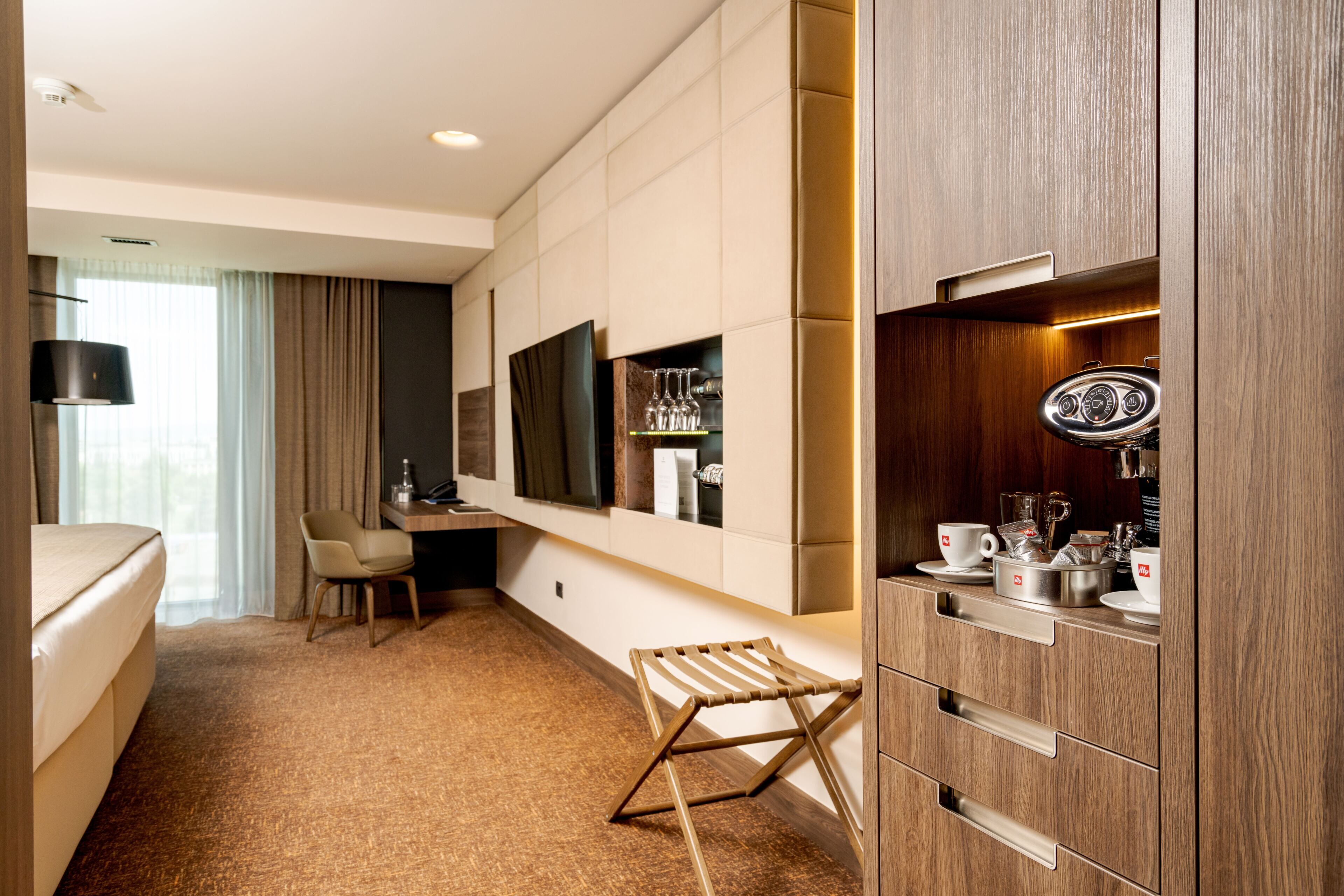 A hotel room with a bed, a desk, a chair, a TV, a minibar, and a window with curtains.