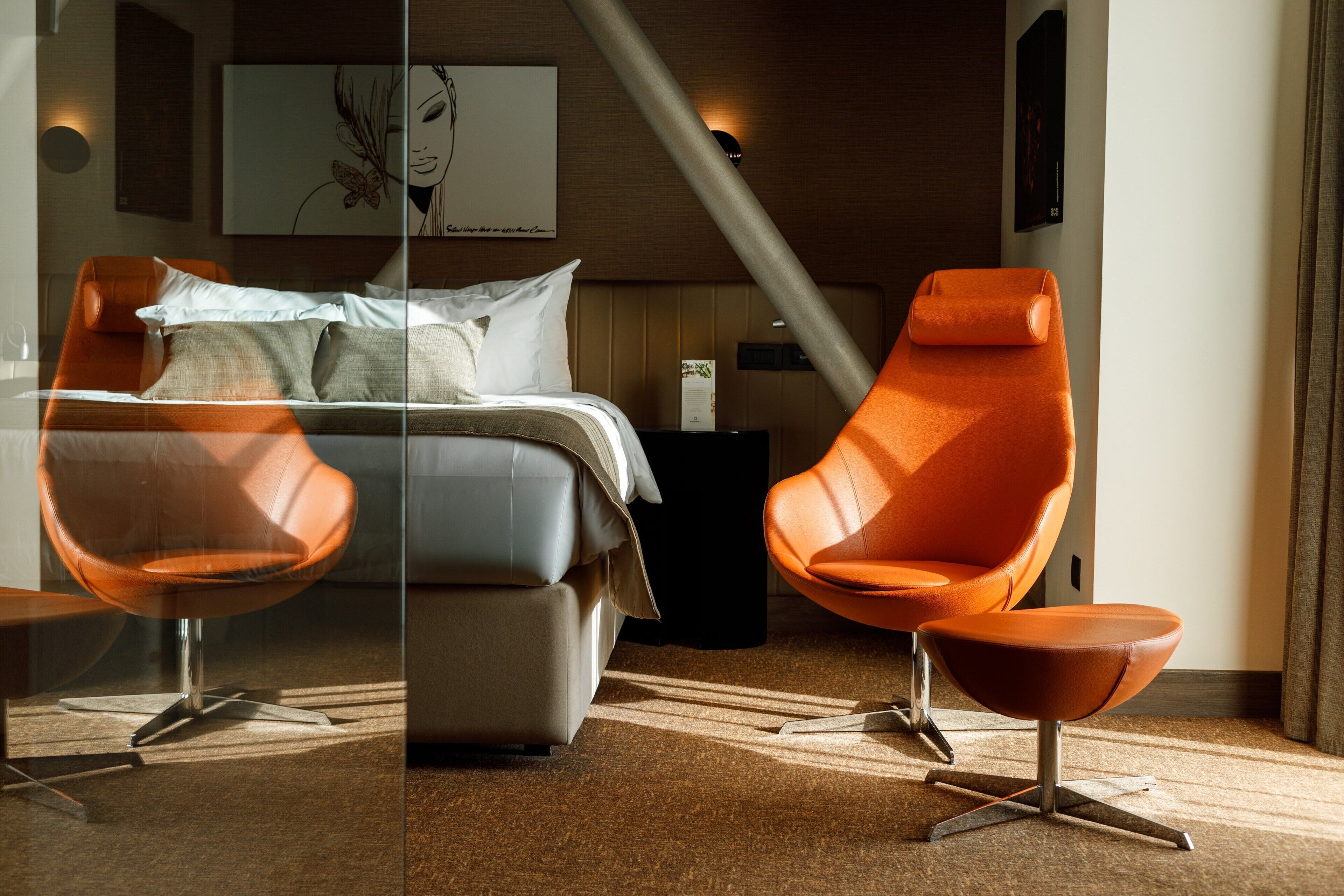 A modern hotel room with a bed, two orange chairs, a glass partition, and a wooden ceiling.