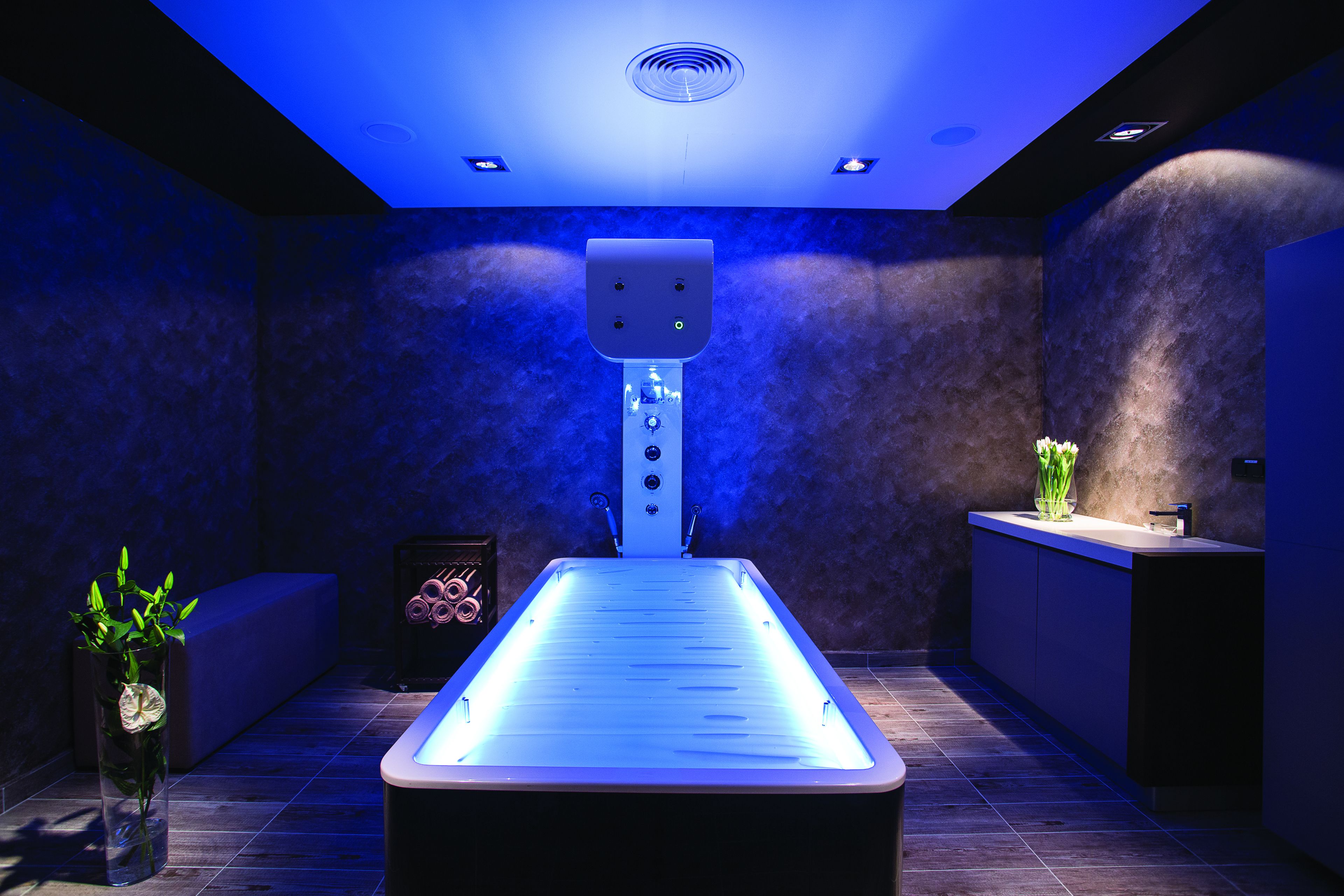 Um spa moderno com banheira de água aquecida, mesa de massagem e um pequeno balcão de atendimento com uma planta.