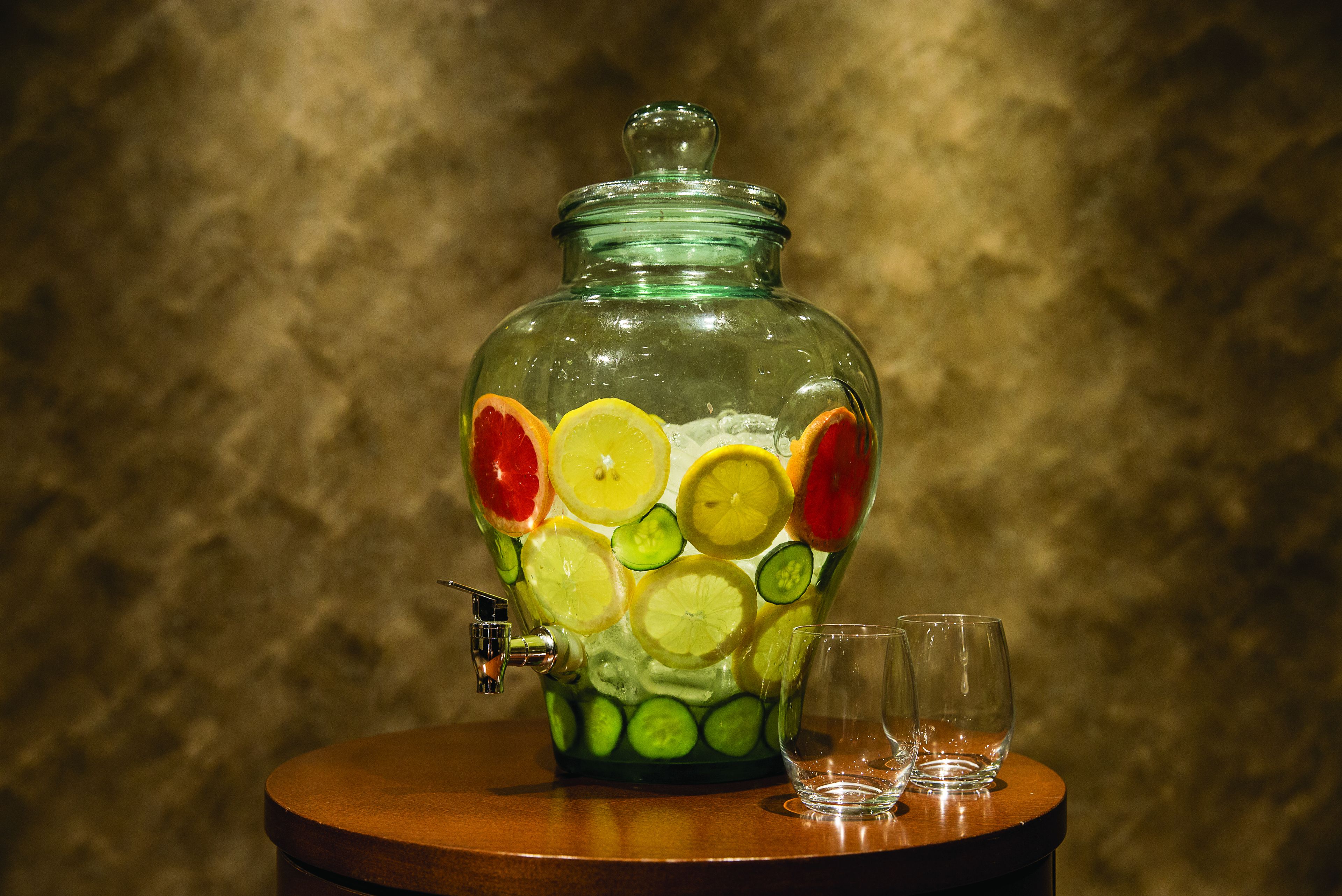 A glass fruit dispenser with a tap, two glasses, and a wooden table.
