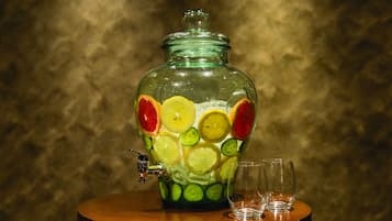A glass fruit dispenser with a tap, two glasses, and a wooden table.