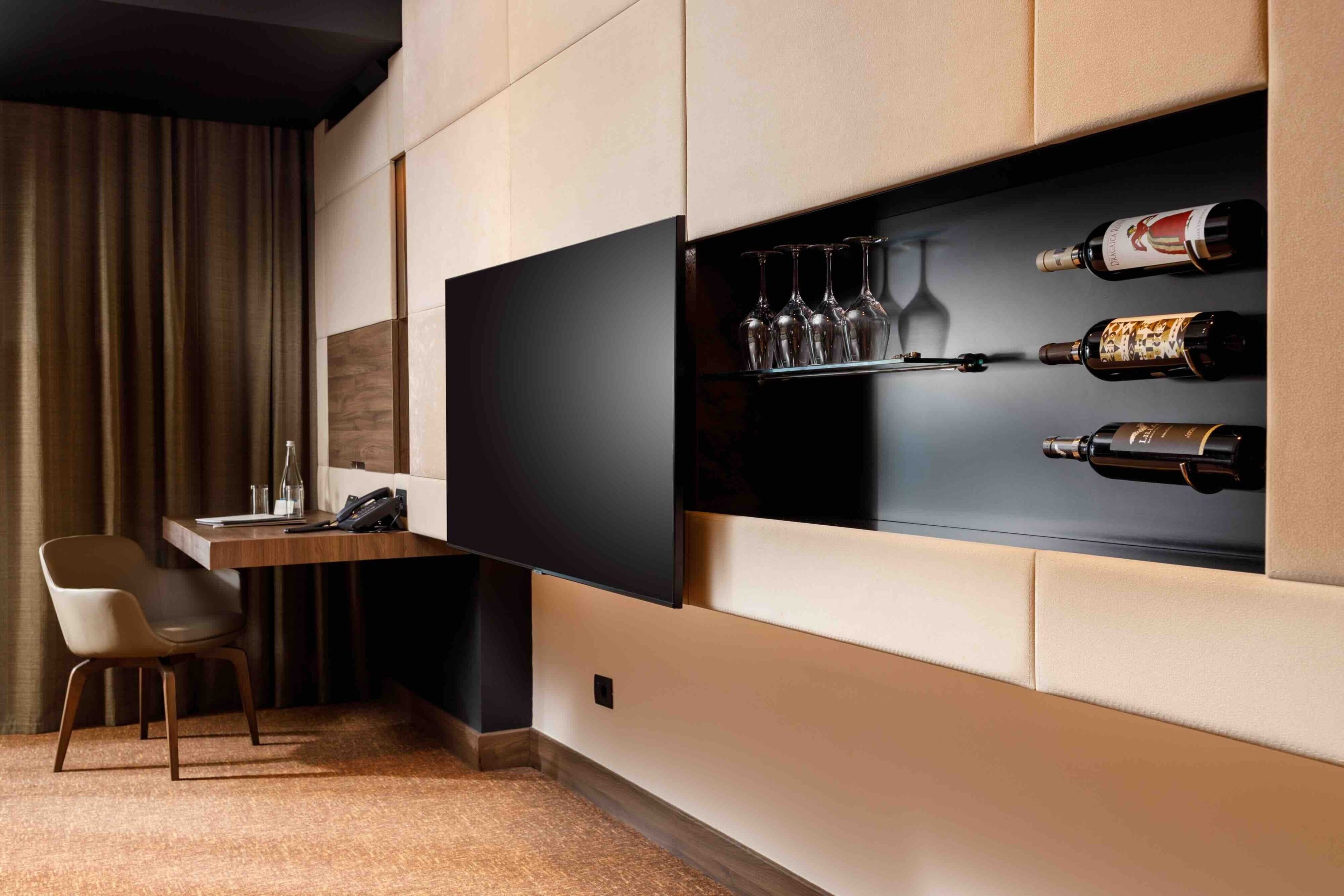 A modern hotel room with a built-in desk, a chair, a wine rack with bottles and glasses, and a large flat-screen TV.