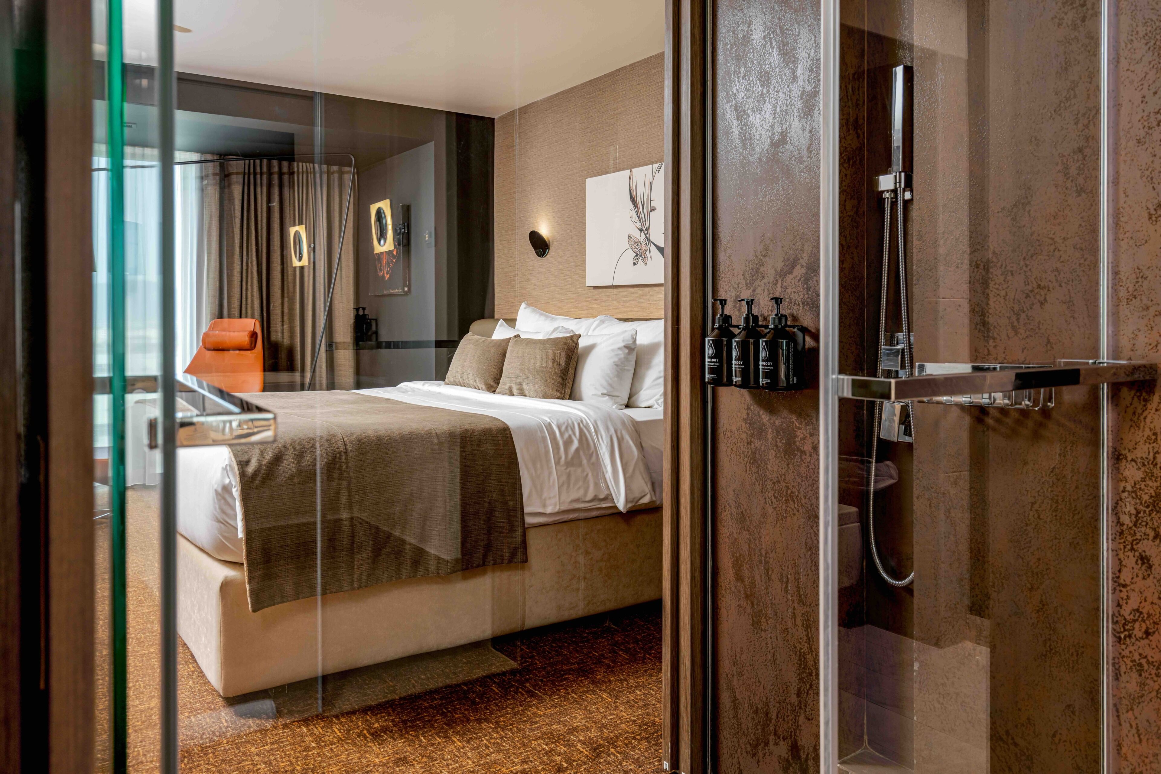 A modern hotel room with a bed, a glass partition, a shower area, and a view of the outside.